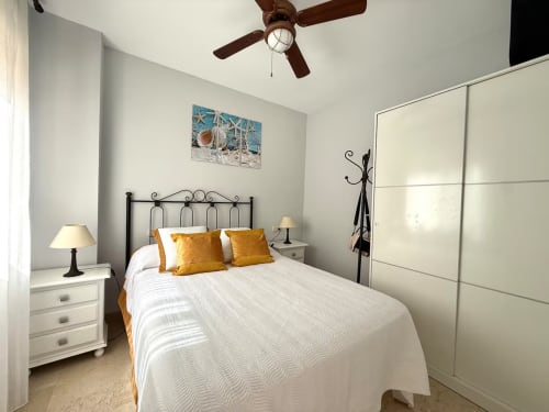 Dormitorio luminoso en apartamento de Fuengirola con ropa de cama blanca, almohadas doradas, estructura metálica, ventilador de techo y armario moderno.