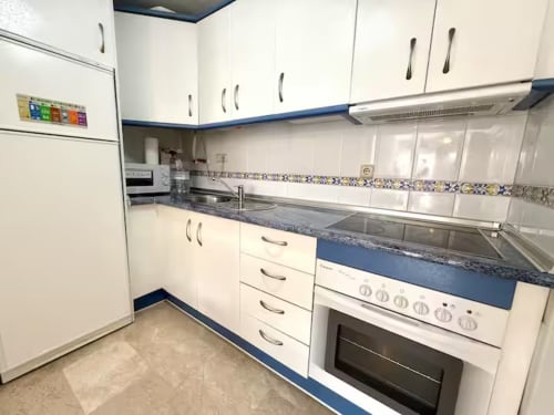 Cocina moderna con muebles blancos, acentos azules, encimera de granito y electrodomésticos de acero inoxidable en apartamento de Fuengirola