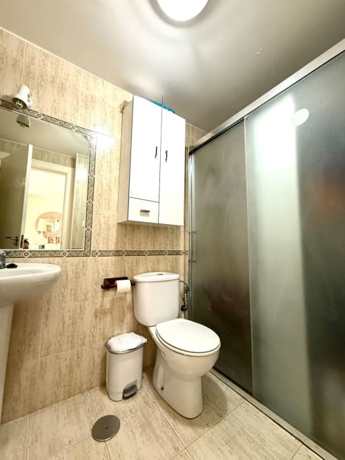 Baño moderno en apartamento de Fuengirola con inodoro blanco, lavabo, espejo y ducha con mampara de cristal y azulejos beige.