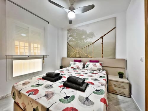 Dormitorio luminoso de apartamento en Fuengirola con ropa de cama estampada, ventilador de techo y cuadro de puente de jungla sobre el cabecero