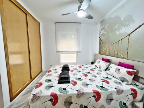 Dormitorio luminoso en apartamento frente al mar en Fuengirola con ropa de cama estampada, ventilador de techo, armario de madera y cuadros de naturaleza