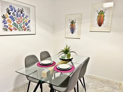 Moderno comedor en apartamento de Fuengirola con mesa de vidrio, sillas grises, obra botánica y escarabajo colorido