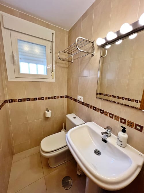 Baño moderno con azulejos beige, lavamanos blanco, inodoro, espejo e iluminación en apartamento de playa en Fuengirola