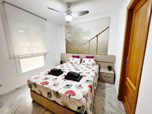Dormitorio moderno en apartamento de playa de Fuengirola con cama matrimonial, ventilador de techo, puerta de madera y ventana con vista a la ciudad