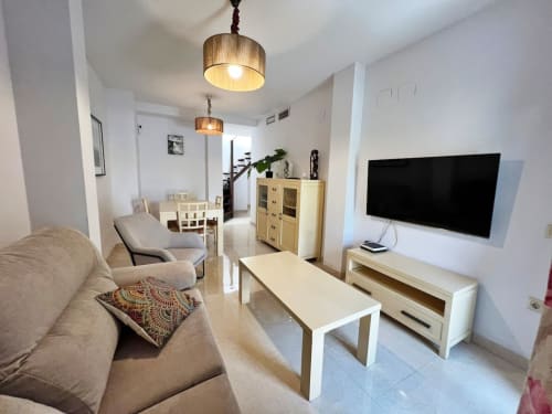 Moderno salón de ático frente al mar en Fuengirola con sofá beige, televisor plano, comedor y mobiliario de madera
