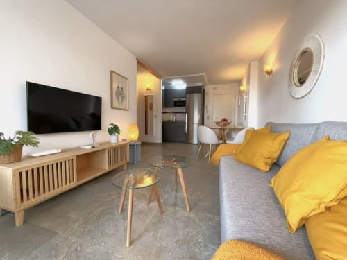 Moderno apartamento soleado en Fuengirola con sofá gris, cojines amarillos, mueble TV de madera y cocina abierta