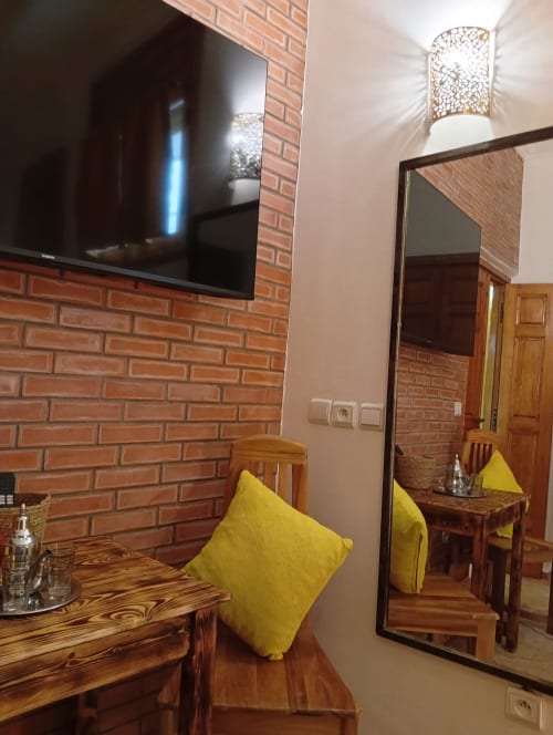 Chambre inspirée par le safran avec coussin jaune, mobilier en bois, mur en brique apparente, TV écran plat et miroir au Riad Le