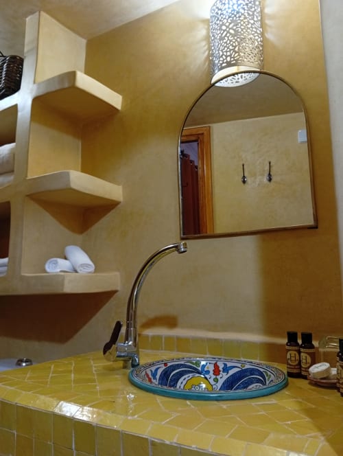 Salle de bain jaune éclatant avec lavabo décoratif, miroir voûté et étagères intégrées au Riad Le Village Mogador, Essaouira