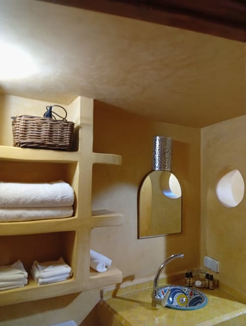 Salle de bains du Riad Le Village Mogador Essaouira, aux murs jaunes, avec étagères intégrées, serviettes, miroir et lavabo