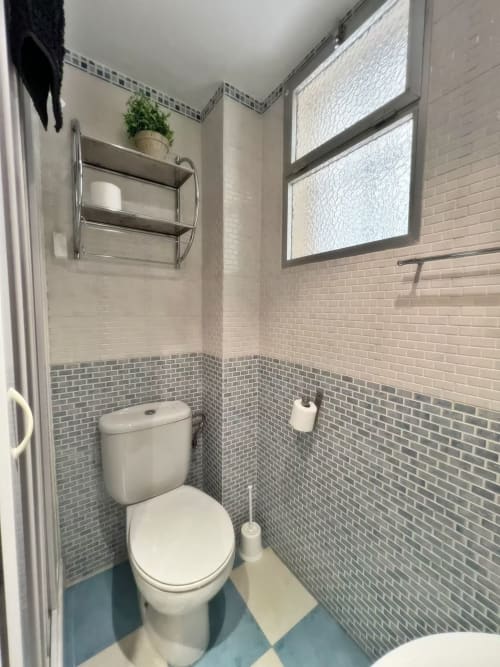Baño moderno en apartamento de Fuengirola con azulejos de ladrillo gris, inodoro blanco, estantería metálica y ventana con vistas a la ciudad