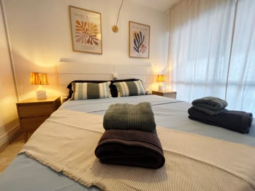 Espacioso dormitorio de apartamento en Fuengirola con decoración moderna, almohadas rayadas, toallas dobladas, mesillas de madera, iluminación cálida,