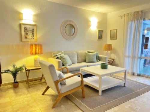 Luminoso salón moderno en apartamento de Fuengirola con sofá crema, butaca de madera y vistas a la ciudad desde la terraza