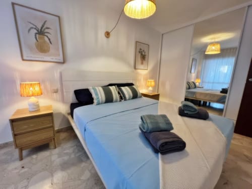 Dormitorio moderno en apartamento de Fuengirola con cama de matrimonio, almohadas rayadas, mesita de noche de madera, espejo e iluminación cálida
