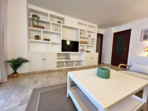 Moderno salón de apartamento en Fuengirola con estantería blanca, televisor, mesa de comedor blanca y vistas a la ciudad