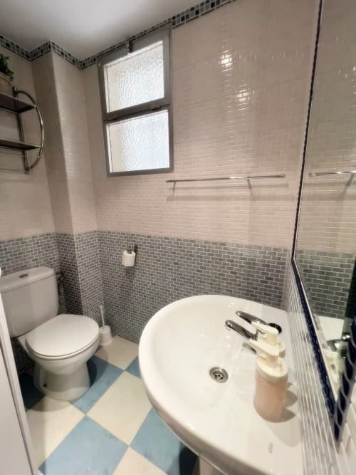 Baño moderno en apartamento de Fuengirola con lavabo blanco, inodoro, azulejos grises y blancos, ventana y ducha con mampara de cristal