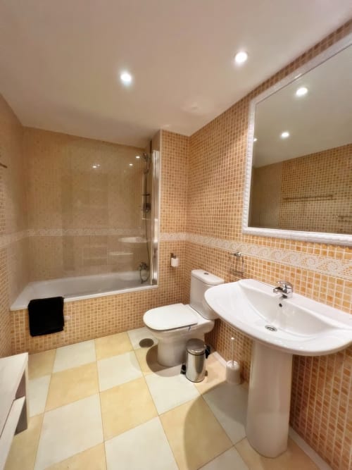 Baño moderno en apartamento de Fuengirola con azulejos de mosaico naranja, lavamanos de pedestal blanco, inodoro y bañera con mampara de cristal,