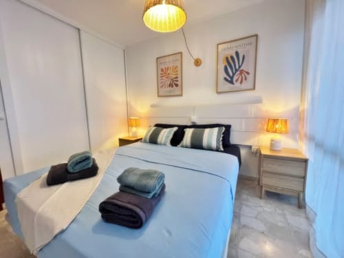 Dormitorio moderno en apartamento de Fuengirola con cama blanca, almohadas rayas, mesitas de noche de madera, iluminación cálida y arte Matisse