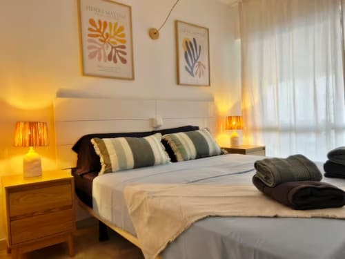 Dormitorio moderno en apartamento de Fuengirola con cabecero blanco, almohadas rayadas, iluminación cálida, obra de Matisse y vistas a la ciudad