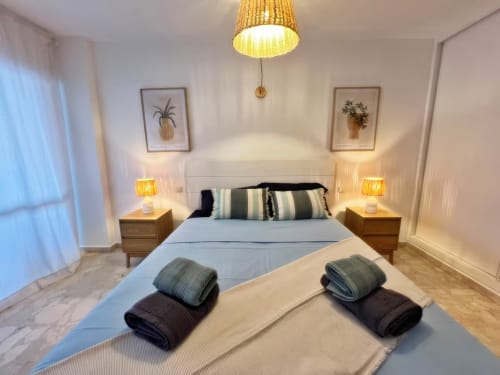 Dormitorio moderno en apartamento de Fuengirola con cabecero blanco, mesillas de madera, almohadas rayadas, toallas enrolladas y lámpara colgante