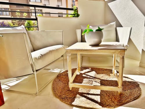 Zona de estar en balcón soleado con silla tapizada color crema, mesa de madera, alfombra tejida y plantas en macetas en Fuengirola