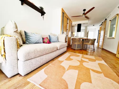 Luminoso salón moderno con sofá crema, alfombra estampada, suelos de madera y ventilador de techo en apartamento playa de Fuengirola