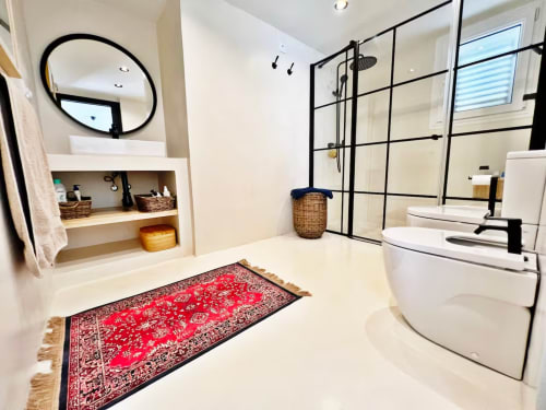 Baño moderno en apartamento de playa en Fuengirola con doble lavabo, ducha a ras de suelo, alfombra roja estampada y espejo circular.