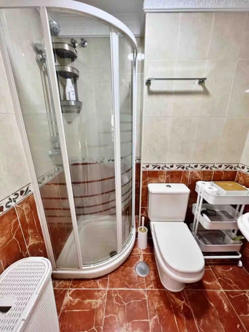 Baño moderno con ducha de cristal curvado, inodoro blanco y azulejos de mármol rojo en apartamento frente al mar en Fuengirola
