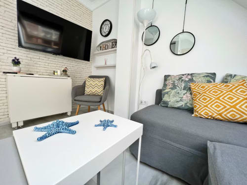 Acogedor apartamento frente al mar en Fuengirola con sofá gris, mesa blanca, TV, reloj y espejos decorativos