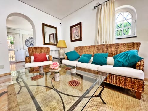 Luminoso salón del apartamento en Fuengirola con muebles de mimbre, cojines turquesas, mesa de cristal y puerta arqueada