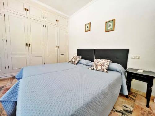 Dormitorio luminoso en apartamento de Fuengirola con ropa de cama azul, cabecero negro, armarios empotrados color crema y suelo de madera