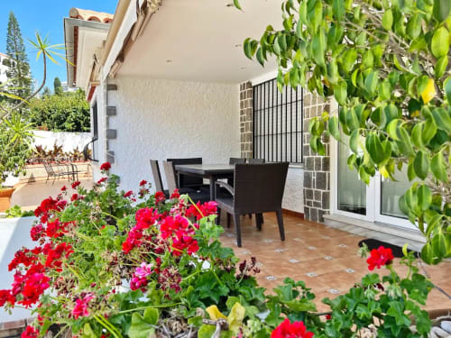 Luminoso apartamento con terraza ajardinada en Fuengirola, geranios rojos, muebles negros y plantas trepadoras verdes