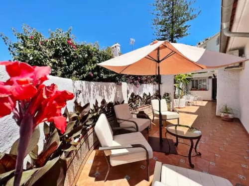 Luminosa terraza con sombrilla naranja, cómodas tumbonas, flores rojas y edificios blancos en apartamento de Fuengirola