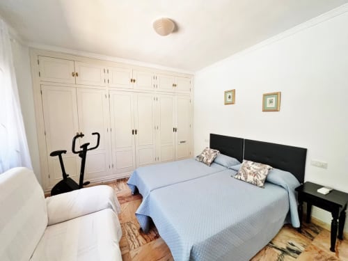 Dormitorio luminoso en apartamento de Fuengirola con cama azul, cabecero negro, armarios empotrados blancos, bicicleta estática y luz natural