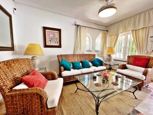 Luminoso salón con muebles de ratán, cojines turquesas, detalles rojo, mesa de cristal y vistas al jardín en apartamento de Fuengirola
