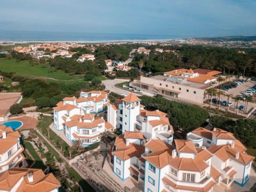 Vista aérea do hotel Be SilverCoast em Serra D'El Rei com edifícios brancos e laranja, jardins, piscina e litoral do oceano