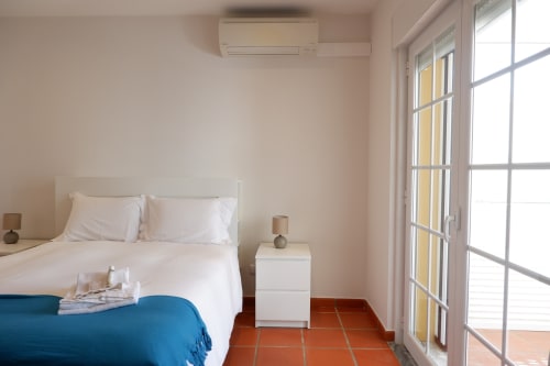 Quarto ensolarado no hotel Be SilverCoast com cama branca, manta azul, pisos em terracota, ar condicionado e portas com vista para o jardim