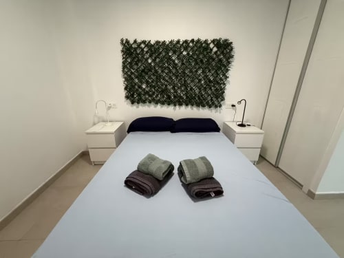 Dormitorio moderno de apartamento en Fuengirola con cama blanca, almohadas oscuras, toallas dobladas y arte botánico en la pared