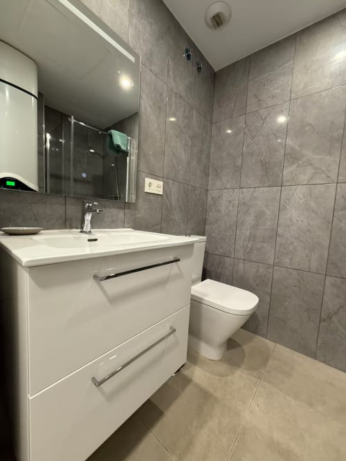 Baño moderno en apartamento frente al mar, Fuengirola. Vanitorios blancos, azulejos grises, inodoro, espejo con iluminación.