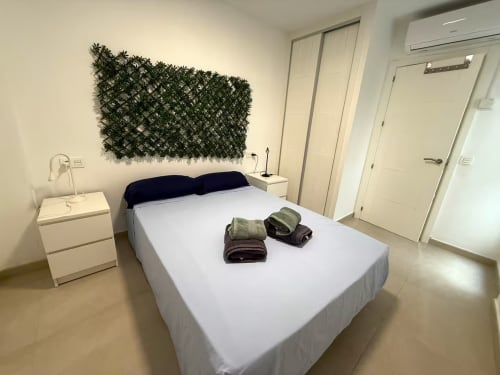 Luminoso dormitorio en apartamento de playa en Fuengirola con cama queen, cabecero oscuro, planta verde, mesita blanca y diseño moderno