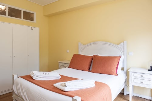 Bright yellow bedroom with white bed, orange pillows, white wardrobe, and neatly folded towels at RH Charming Apartments in