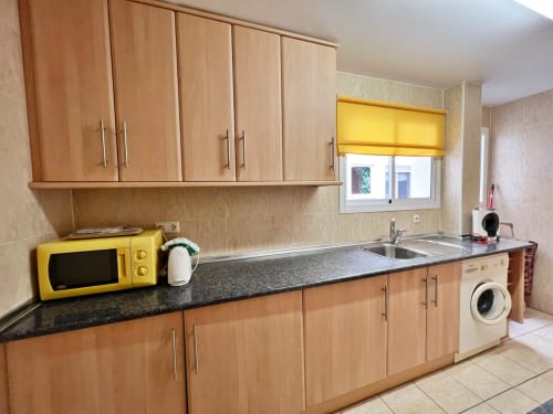 Cocina moderna con gabinetes de madera, encimera oscura, microondas amarillo y lavadora en apartamento frente al mar,