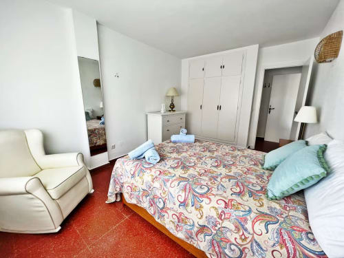 Dormitorio luminoso en apartamento frente al mar en Fuengirola con ropa de cama estampada, armario blanco, sillón y vistas al mar
