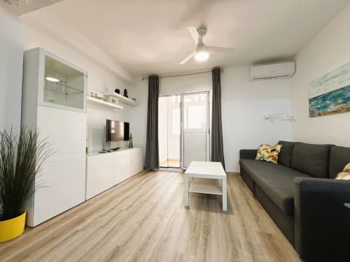 Moderna sala de estar en Fuengirola con sofá oscuro, cocina blanca, televisión y acceso a balcón