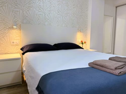 Dormitorio moderno en apartamento de Fuengirola con ropa de cama blanca, almohadas negras, patrón geométrico en pared, iluminación cálida y doblado
