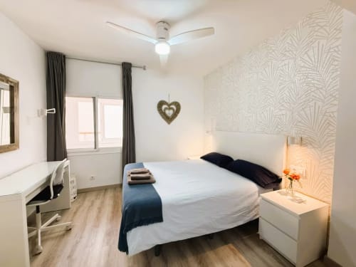 Dormitorio moderno en apartamento de Fuengirola con cama de matrimonio, ventilador de techo, mesita blanca, escritorio y papel tapiz estampado