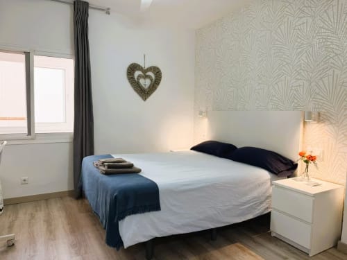 Moderno dormitorio en apartamento de Fuengirola con cama de matrimonio, cabecero blanco, almohadas negras, mesita de noche y decoración de corazón