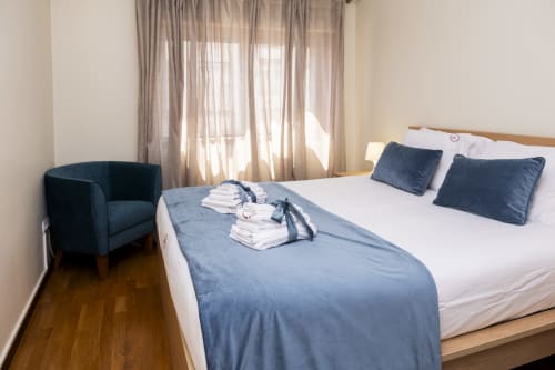 Spacious bedroom at RH Charming Apartments in Barcelos with queen bed, navy pillows, blue armchair, and city view from window