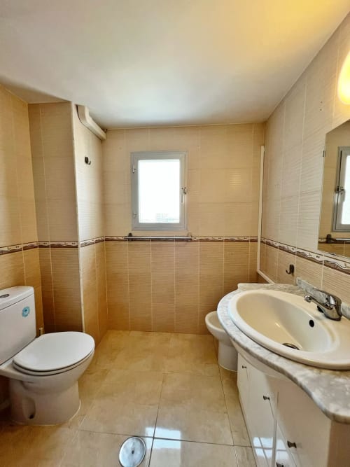 Moderno baño en apartamento frente al mar de Grupo Inmo House, Fuengirola, con paredes de azulejos beige, lavabo, inodoro y ventana.