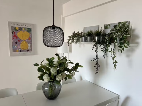 Interior de apartamento luminoso con paredes blancas, lámpara colgante, plantas en maceta en estante y flores frescas en jarrón de cristal
