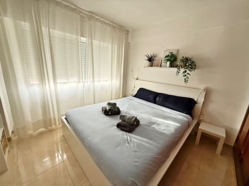 Dormitorio luminoso de apartamento en Fuengirola con sábanas blancas, almohadas negras, plantas en maceta en estantería y cortinas translúcidas